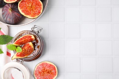 Glass jar of tasty sweet fig jam and fruits on white tiled table, flat lay. Space for text