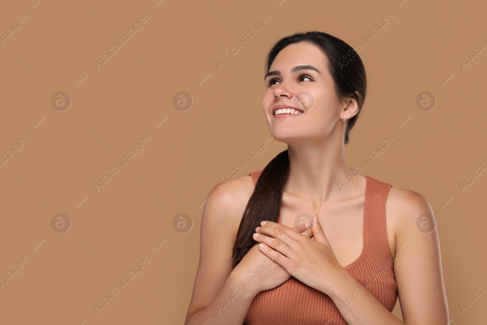 Photo of Thank you gesture. Beautiful grateful woman holding hands near heart on brown background, space for text