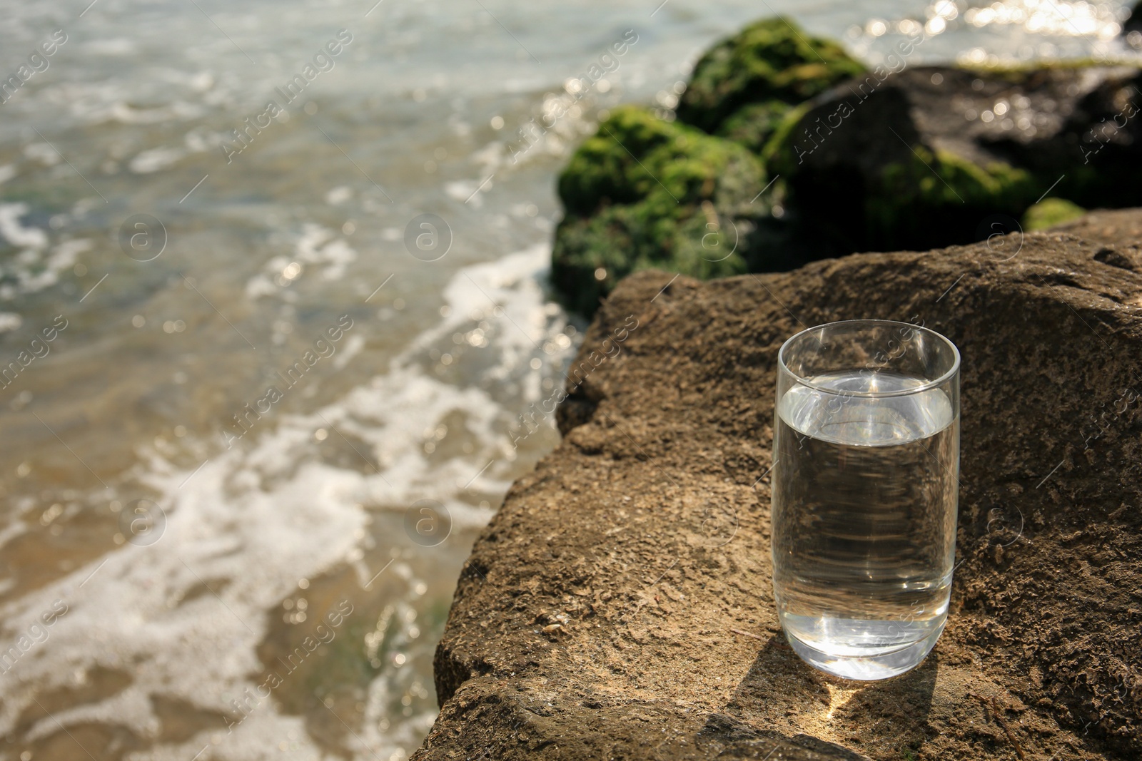 Photo of Glass of fresh water on stone near sea, space for text