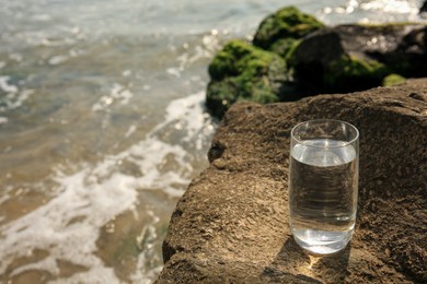 Glass of fresh water on stone near sea, space for text