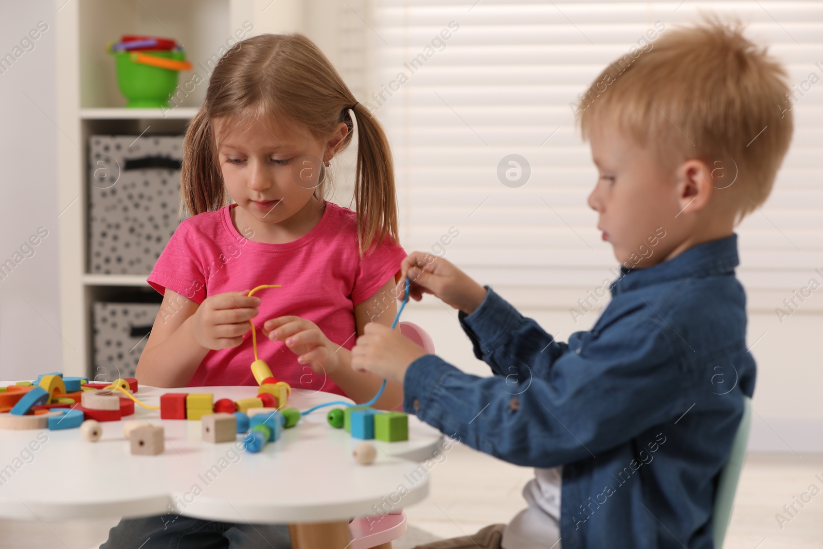 Photo of Little children playing with wooden pieces and string for threading activity at white table indoors. Developmental toys