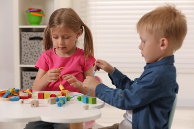 Little children playing with wooden pieces and string for threading activity at white table indoors. Developmental toys