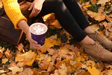 Photo of Woman holding cup of hot drink in park with fallen leaves, closeup view. Autumn season