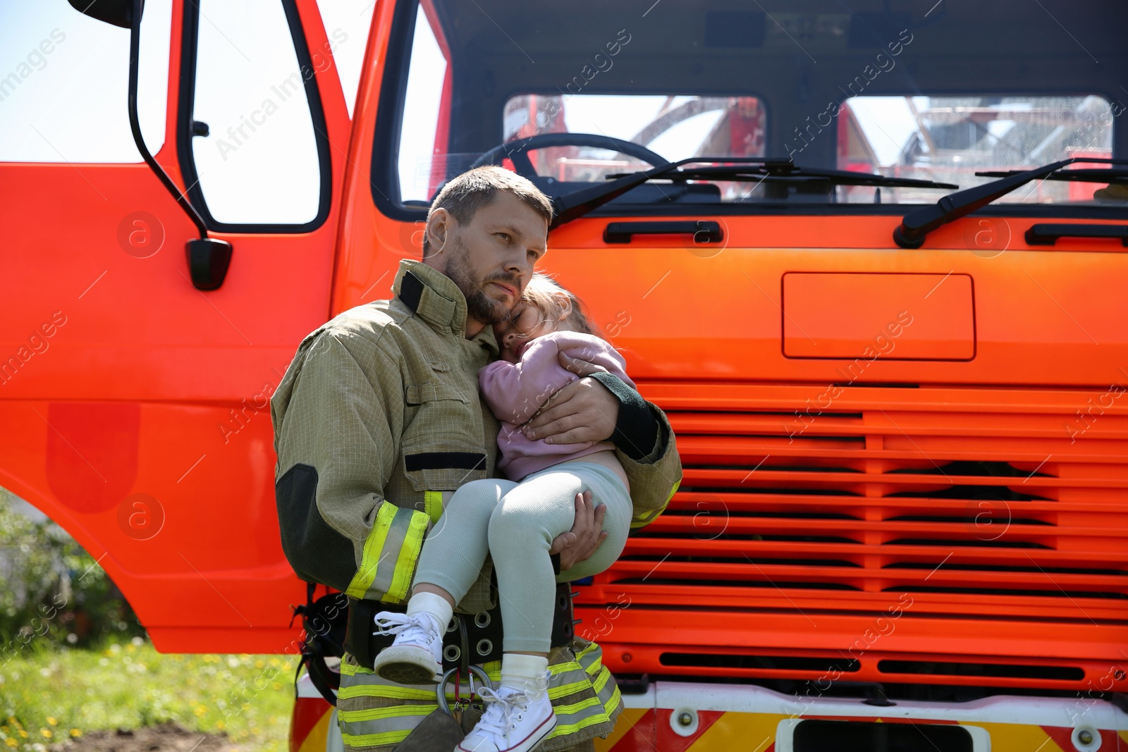 Photo of Firefighter in uniform holding rescued little girl near fire truck outdoors. Save life