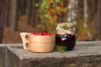 Photo of Tasty lingonberry jam and cup with red berries on wooden table in forest