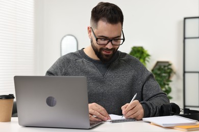 Photo of E-learning. Young man taking notes during online lesson at white table indoors