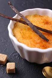 Photo of Delicious creme brulee in bowl, vanilla pods and sugar cubes on dark gray textured table, closeup