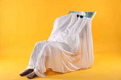 Photo of Person in ghost costume and sunglasses relaxing on deckchair against yellow background