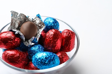 Photo of Glass bowl with chocolate eggs wrapped in colorful foil on white background, closeup