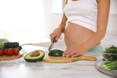 Photo of Young pregnant woman cutting avocado at table in kitchen, closeup. Taking care of baby health
