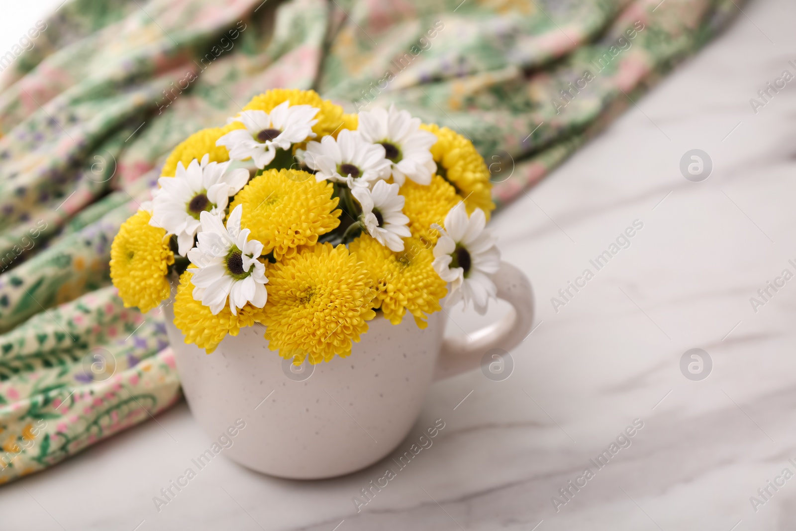 Photo of Beautiful bright flowers in cup and fabric on white marble table, closeup