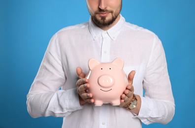 Young man with piggy bank on light blue background, closeup