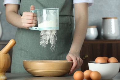 Photo of Woman sieving flour into bowl at table in kitchen, closeup