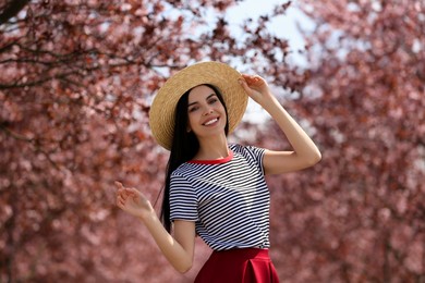 Photo of Pretty young woman with straw hat near beautiful blossoming trees outdoors. Stylish spring look