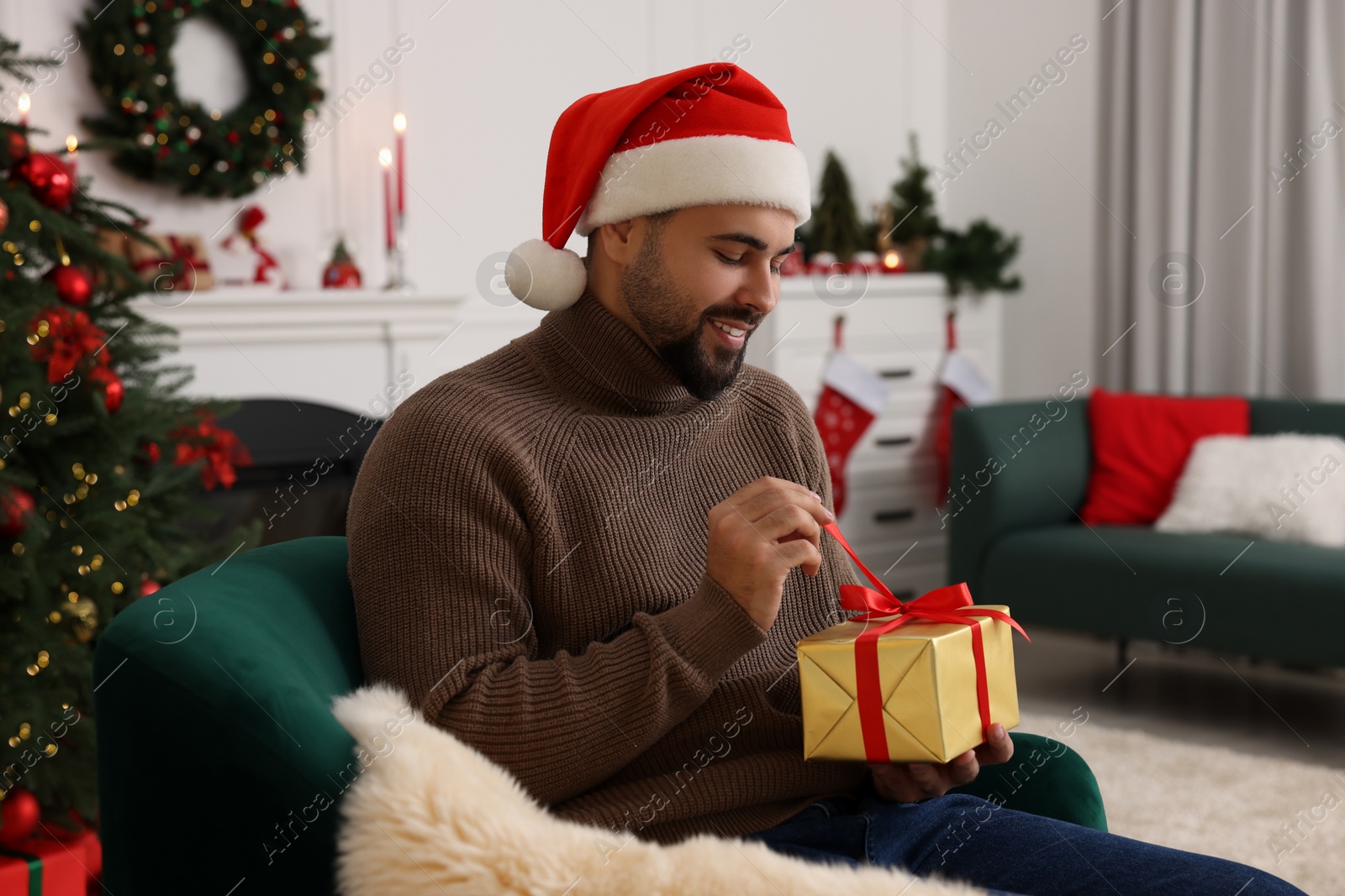 Photo of Happy young man in Santa hat opening Christmas gift at home