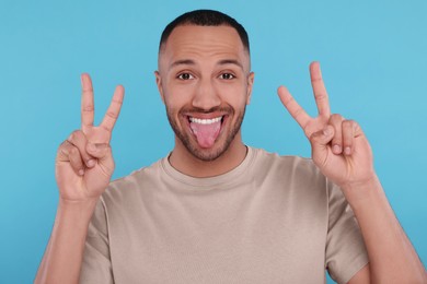 Photo of Happy young man showing his tongue and V-sign on light blue background