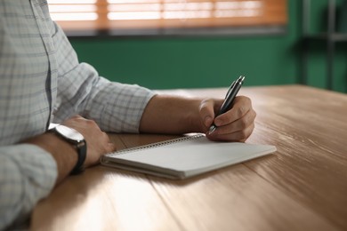 Left-handed man writing in notebook at wooden table indoors, closeup