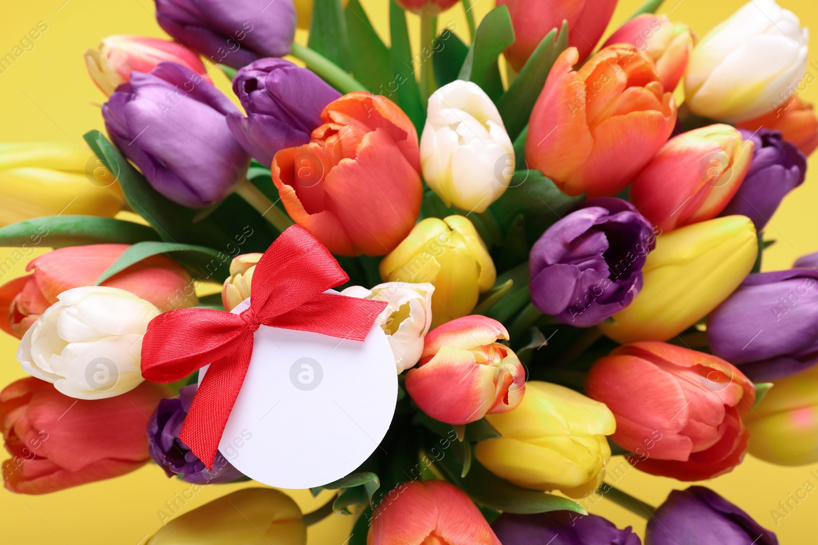 Photo of Bouquet of beautiful colorful tulips with blank card on yellow background, closeup. Birthday celebration
