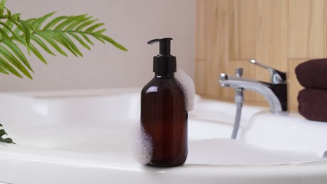 Photo of Bottle of bubble bath with foam on tub indoors