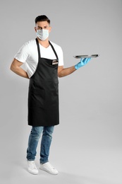 Waiter in medical face mask with empty tray on light grey background