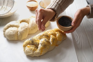 Photo of Woman adding poppy seeds onto braided dough at white wooden table in kitchen, closeup. Cooking traditional Shabbat challah