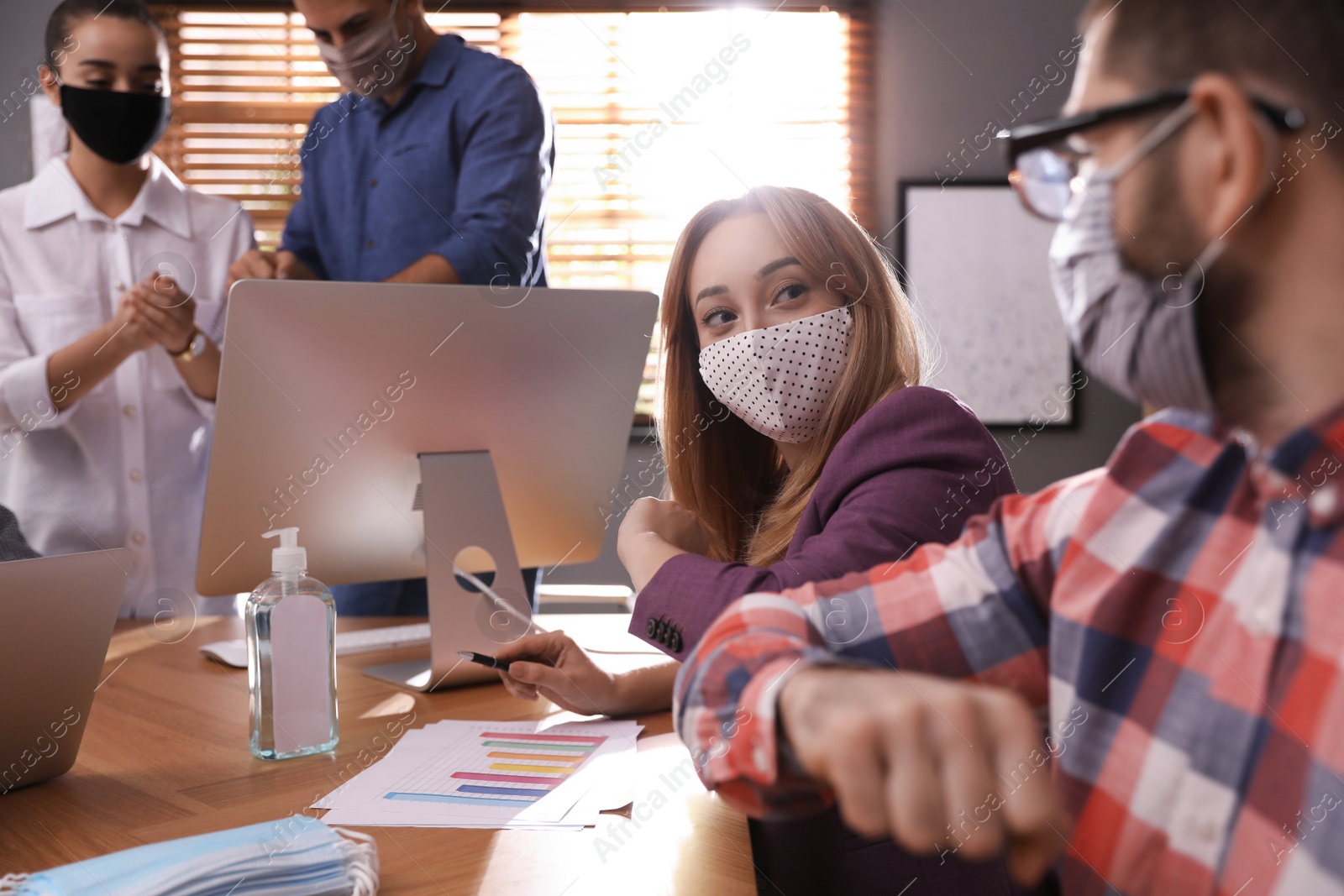 Photo of Coworkers with protective masks making elbow bump in office. Informal greeting during COVID-19 pandemic