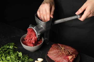 Woman making beef mince with manual meat grinder at dark textured table against black background, closeup