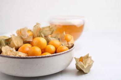 Photo of Ripe physalis fruits with calyxes in bowl on white tiled table, closeup. Space for text