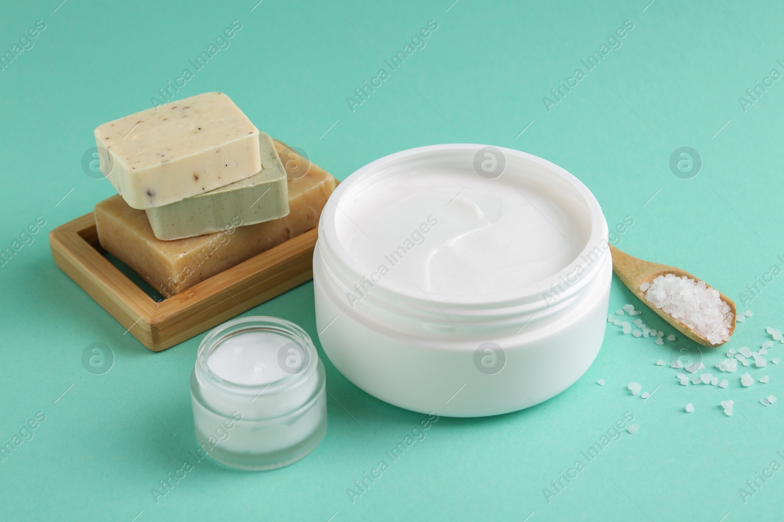 Photo of Moisturizing cream in open jars, sea salt and soap bars on turquoise background. Body care products