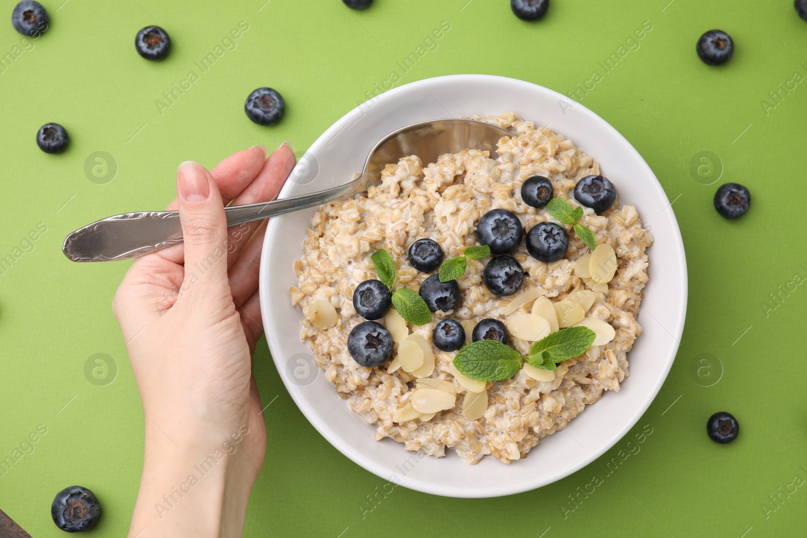 Photo of Woman eating tasty oatmeal with blueberries, mint and almond petals on light green background, top view