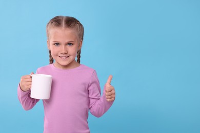 Photo of Happy girl with white ceramic mug showing thumbs up on light blue background, space for text