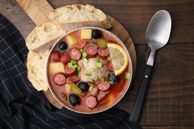 Photo of Meat solyanka soup with thin dry smoked sausages in bowl and bread slices on wooden table, flat lay