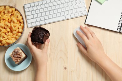 Photo of Bad habits. Woman eating muffin while working on computer at wooden table, top view