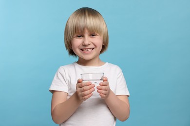 Happy little boy holding glass of fresh water on light blue background