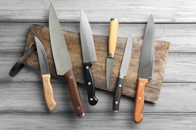 Photo of Many different knives and board on light wooden table, flat lay