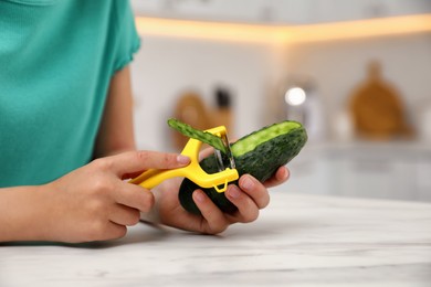 Photo of Little girl peeling cucumber at table in kitchen, closeup. Preparing vegetable