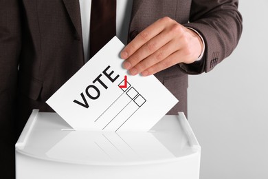 Man putting paper with word Vote and tick into ballot box on light grey background