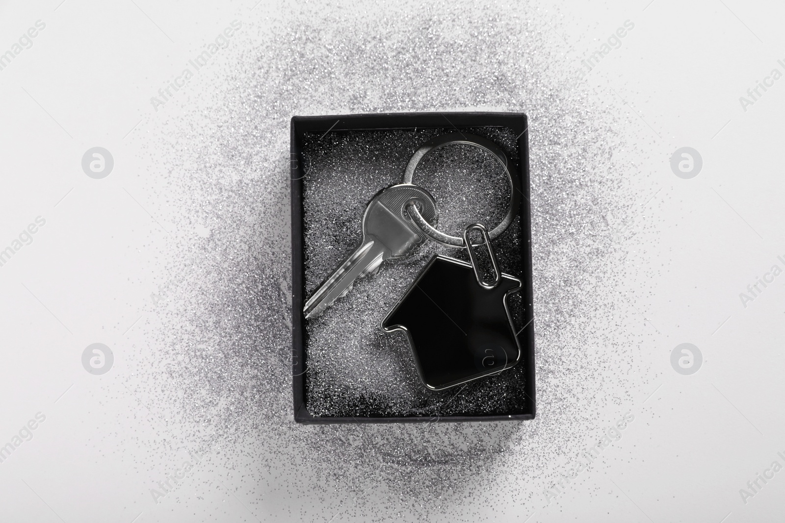 Photo of Key with trinket in shape of house, glitter and gift box on light grey background, top view. Housewarming party