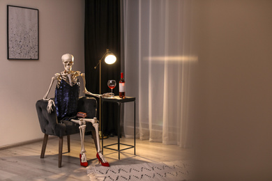 Photo of Skeleton in dress with wine sitting at table indoors