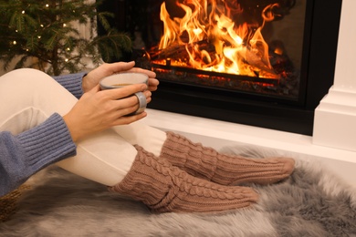 Photo of Woman with cup of drink sitting near burning fireplace at home, closeup