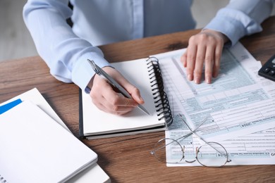 Photo of Payroll. Woman taking notes in notebook while working with tax return forms at wooden table, closeup