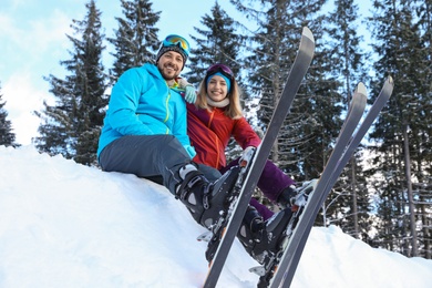 Happy couple with ski equipment sitting on snowdrift outdoors. Winter vacation