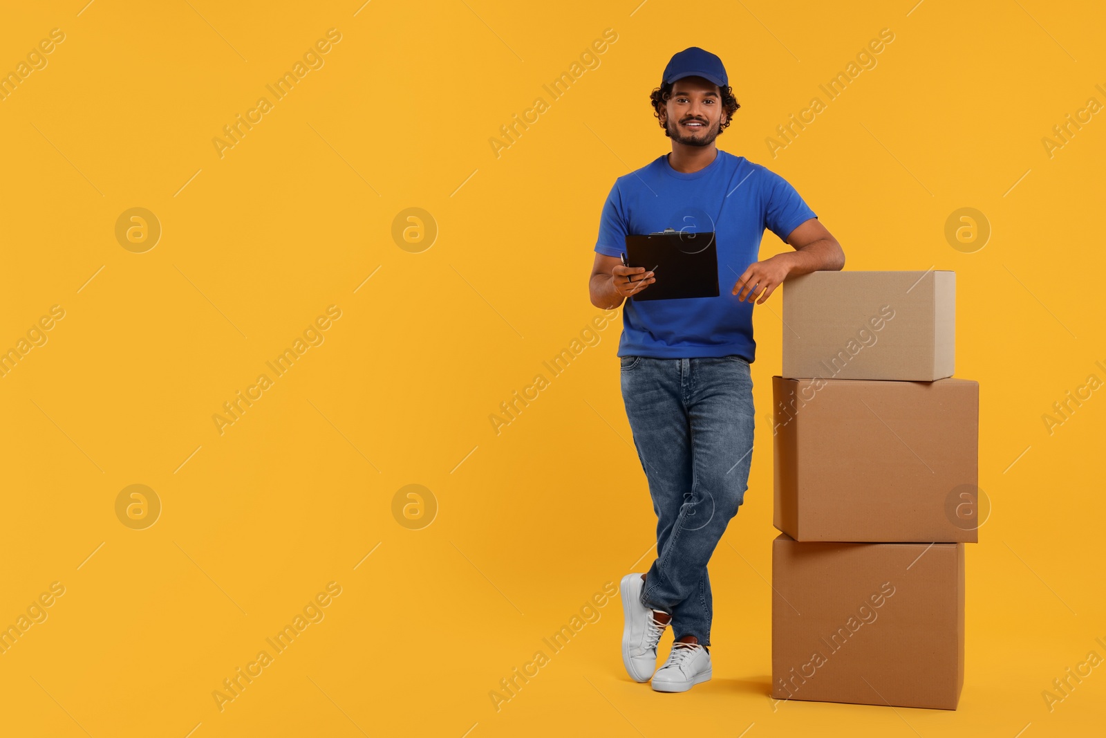 Photo of Happy courier with clipboard and stack of parcels on orange background, space for text