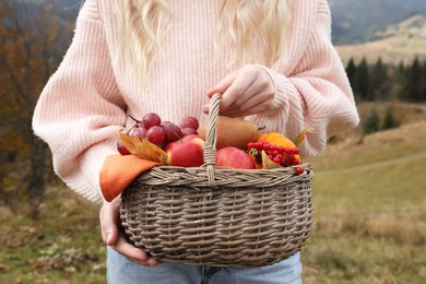 Photo of Woman holding wicker picnic basket with fruits and autumn leaves outdoors, closeup