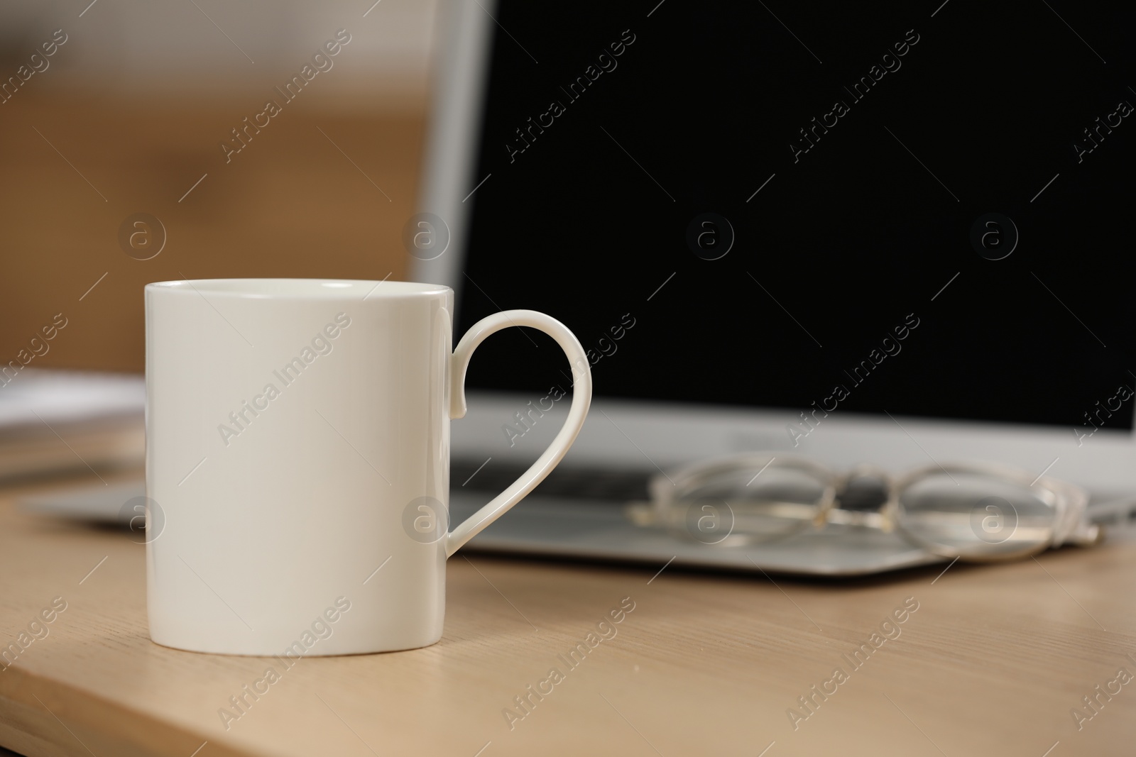 Photo of White ceramic mug and laptop on wooden table at workplace. Space for text