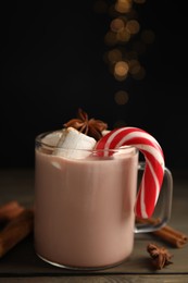 Photo of Glass cup of tasty cocoa with marshmallows and Christmas candy cane on wooden table against blurred festive lights
