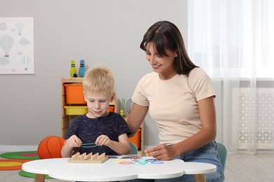 Photo of Motor skills development. Happy mother helping her son to play with geoboard and rubber bands at white table in room