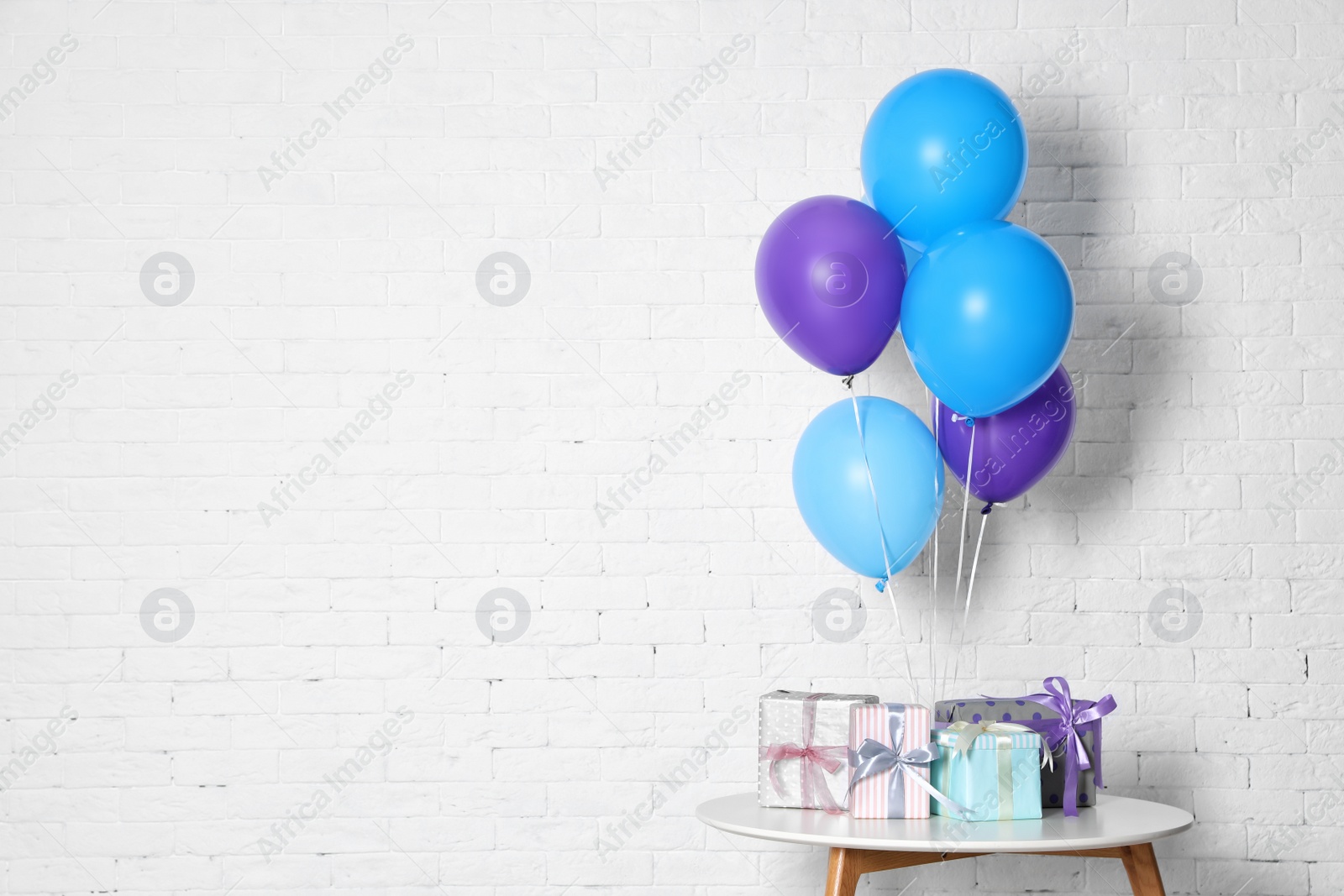 Photo of Bunch of bright balloons and gifts on table against white brick wall, space for text