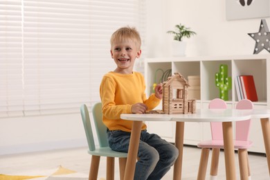 Cute little boy playing with wooden house at white table indoors, space for text. Child's toy