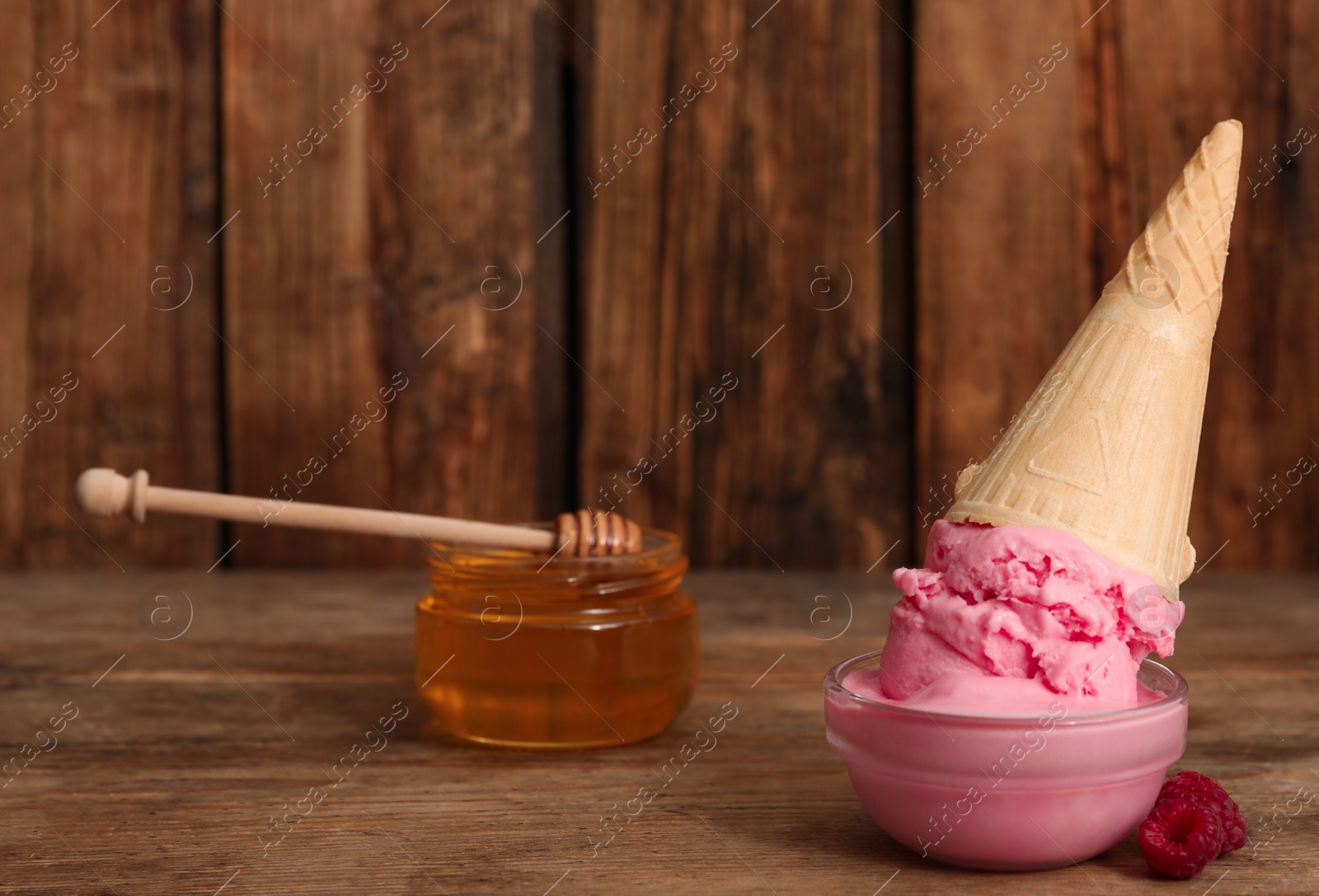 Photo of Delicious pink ice cream in wafer cone, honey and raspberries served on wooden table. Space for text